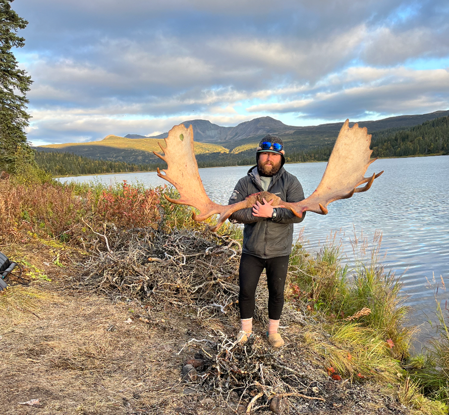 Person holding large moose antlers by a lakeside amid scenic mountains.